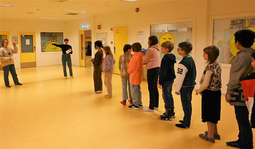 Bildet viser en gruppe barn stående på en rekke, med en voksen i front som peker til høyre. - Klikk for stort bilde
